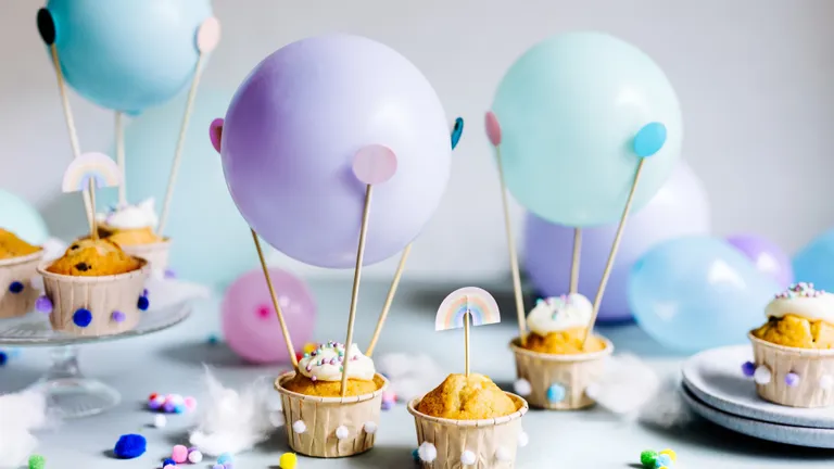 Cupcakes mit Zuckerguss und Streuseln, verziert mit pastellfarbenen Luftballons und Regenbogen-Dekorationen, umgeben von bunten Pompons vor einem hellen Hintergrund.