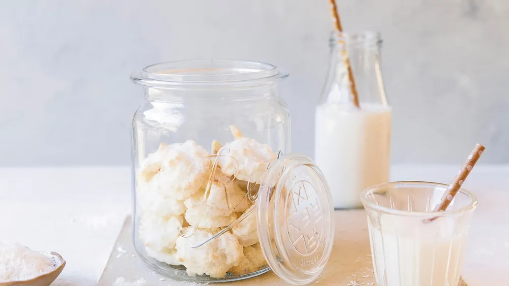 Ein Glas mit Kokosmakronen neben zwei Gläsern Milch mit Papierstrohhalmen vor einem hellen Hintergrund.