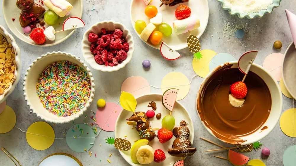 Auf einem Tisch stehen verschiedene Obstsorten, Marshmallows und Toppings sowie ein Schokoladenfondue, umgeben von bunten Papierdekorationen.