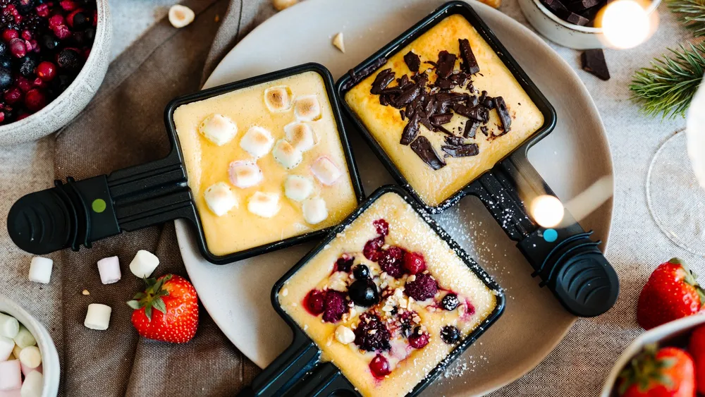 Drei kleine Auflaufförmchen mit gebackenen Desserts, garniert mit Marshmallows, Schokoladenraspeln und gemischten Beeren, umgeben von Erdbeeren und Schalen.