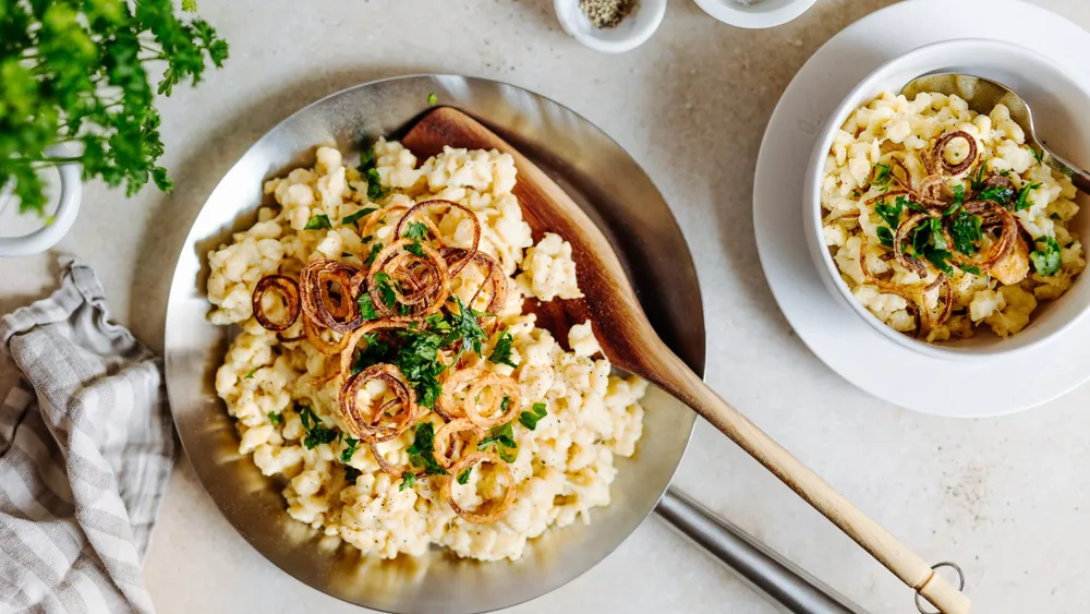 Käsespätzle in einer silbernen Pfanne mit Holz-Kochlöffel auf hellem Hintergrund