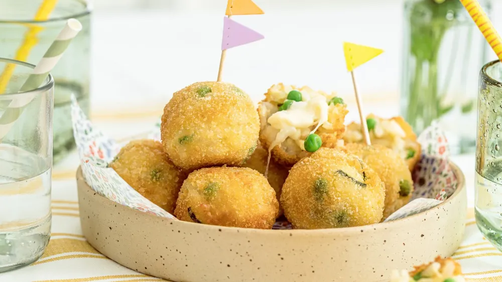 Goldbraun gebratene Arancini mit Käse und Erbsen, garniert mit kleinen Fähnchen, in einer gesprenkelten Schüssel auf einer gestreiften Tischdecke.