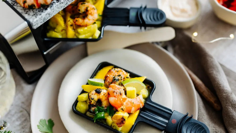 Gegrillte Garnelen mit Mangowürfeln und Limettenscheiben in einer kleinen schwarzen Pfanne, servierfertig auf einem weißen Teller, daneben eine Dip-Sauce.