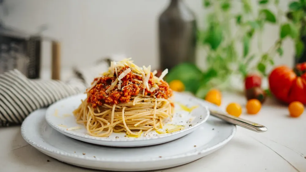 Ein Teller Spaghetti mit Tomatensoße und geriebenem Käse, garniert mit schwarzem Pfeffer, auf einem rustikalen Tisch mit bunten Tomaten im Hintergrund.