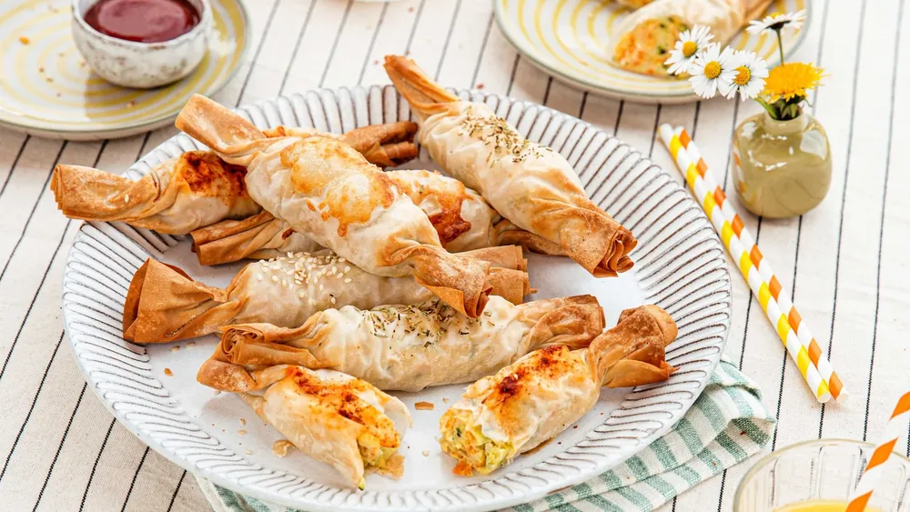 Ein Teller mit knusprigen Blätterteigröllchen, garniert mit Sesam und Kräutern, dazu eine Dip-Sauce, gestreifte Strohhalme und Blumen im Hintergrund.