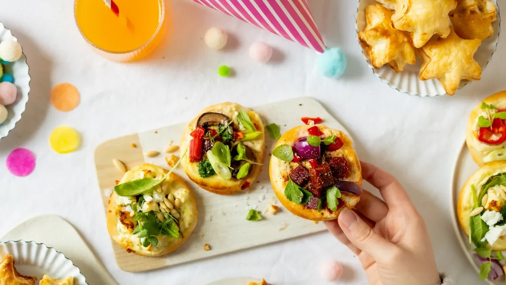 3 Mini Pizzen auf einem Jausenbrettchen, darunter ein bunt dekorierter Tosch mit weißer Tischdecke