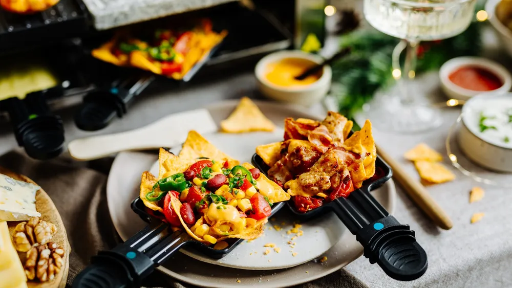 Zwei Raclettepfännchen mit reichhaltig belegten Nachos: einer mit Käse, Bohnen und Jalapeños, der andere mit Hähnchen und Käse, dazu verschiedene Dips und Beilagen.