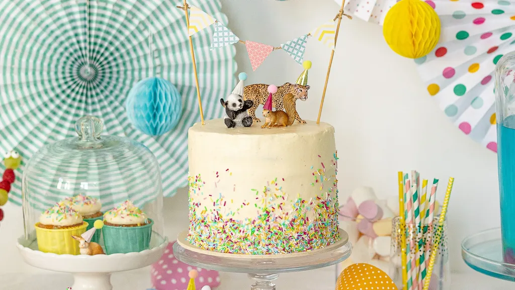 Geburtstagstorte mit Tierfiguren, bunten Streuseln und Mini-Girlanden. Dazu gehören Cupcakes und gestreifte Papierstrohhalme.