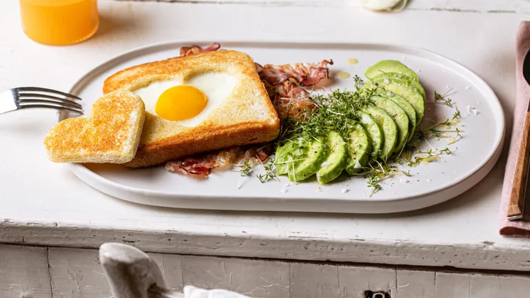 herzförmige Eier mit gebratenem Speck, Toast und Avocado auf einem hellen Tisch