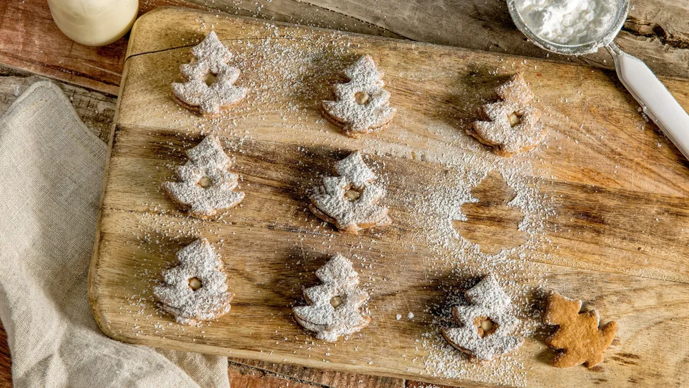 Baumförmige Kekse, mit Puderzucker bestäubt, auf einem Holzbrett, daneben eine weiße Serviette und ein Sieb