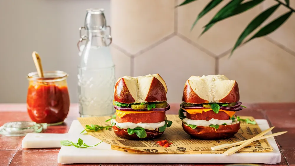 Veganer Leberkäse Burger auf einem Schneidebrett angerichtet, daneben eine Bügelflasche und ein Glas mit Relish