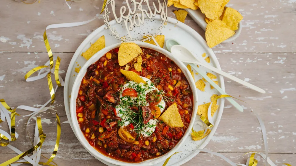 Eine Portion Eintopf garniert mit Sauerrahm und TOrtilla Chips in einer weißen Porzellanschüssel, rundherum Faschingsdeko auf einem hellen Tisch