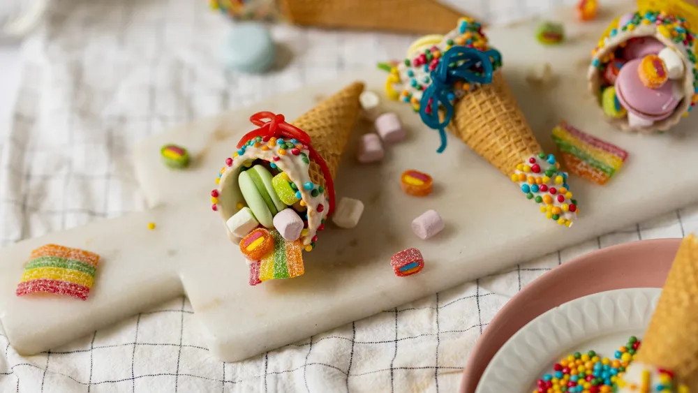 Bunte, mit Süßigkeiten gefüllte Waffeltüten mit Streuseln und Marshmallows auf einem Marmorbrett, das auf einer karierten Tischdecke steht.