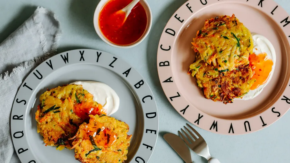 Zwei Teller mit Gemüsefrikadellen, Sauerrahm und Chilisauce. Die Teller sind mit Buchstaben verziert und liegen neben einer Serviette, einer Gabel, einem Messer und einer Sauciere.