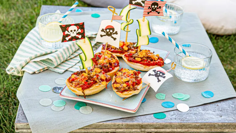 Mini-Pizzen mit Piratenflaggen auf einem Tablett, umgeben von Papierkonfetti und Getränken mit Zitronenscheiben auf einem Tisch im Freien.