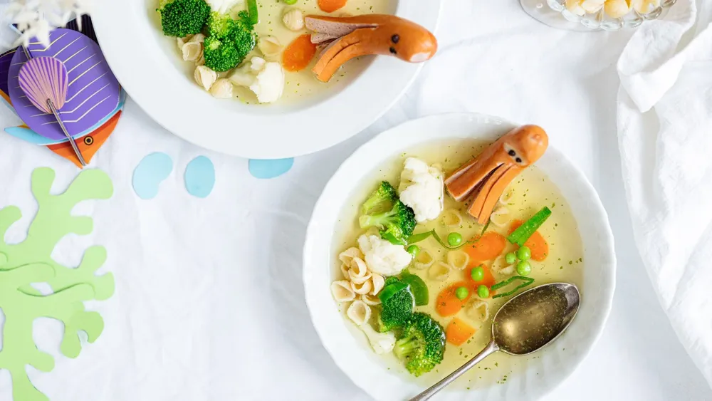 Zwei Schüsseln Gemüsesuppe mit Würstchen in Form von Tintenfischen, gefüllt mit Brokkoli, Blumenkohl, Erbsen, Karotten und Muschelnudeln.