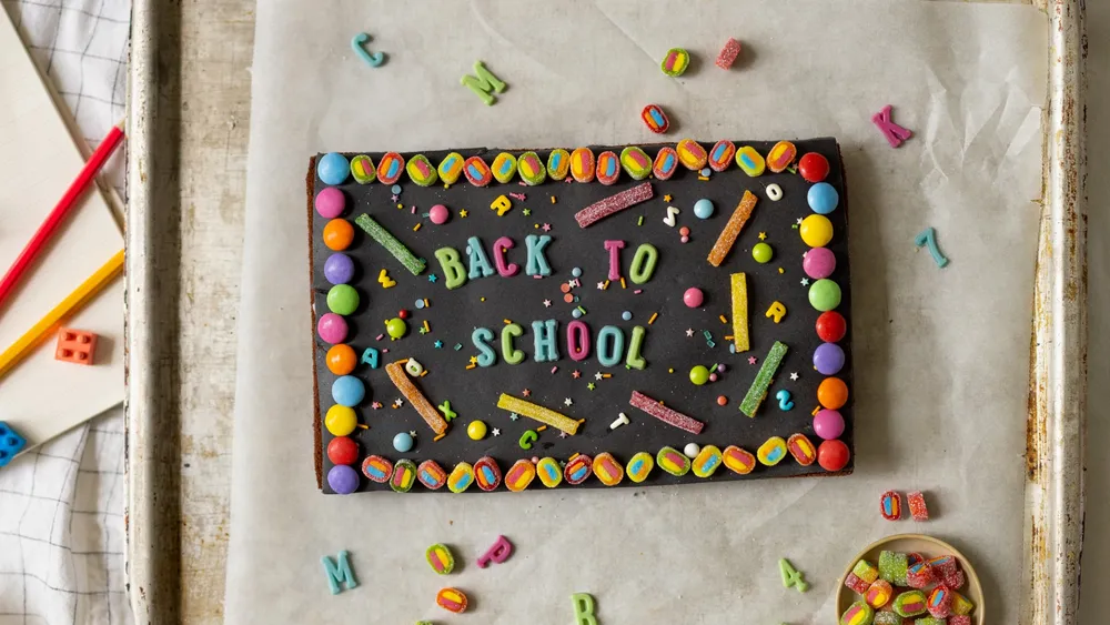 Ein bunter Schokoladenkuchen mit Süßigkeitenverzierungen auf Backpapier, umgeben von Gegenständen zum Thema Schule, verkündet: „Schulbeginn“.