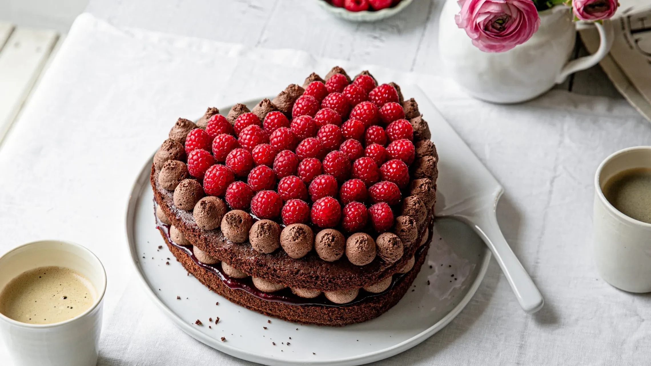 Herzförmige Schokotorte mit Himbeeren auf einem hellen Tisch mit Kaffeetassen