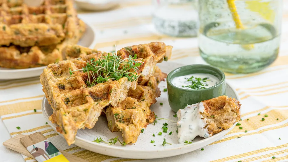 Herzhafte Waffeln mit Sprossen, serviert mit einer grünen Dip-Sauce, auf einem weißen Teller vor einem gestreiften Stoffhintergrund.