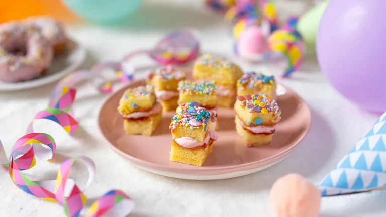 Mini sponge cakes with pink icing and sprinkles on a plate, surrounded by colorful decorations and balloons.