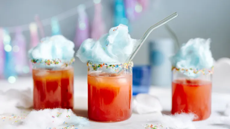 Drei bunte Getränke mit orangeroter Flüssigkeit, garniert mit blauer Zuckerwatte und bunten Streuseln, jeweils mit einem Strohhalm, auf einem festlich gedeckten Tisch.