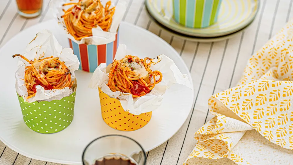 Spaghetti-Nester mit Käse und Tomatensoße in bunten Pappbechern auf einem weißen Teller, serviert auf einer gestreiften Tischdecke mit einer gemusterten Serviette.