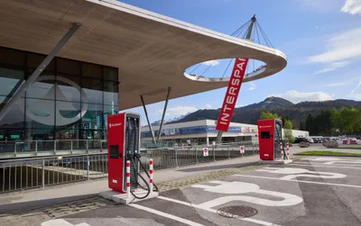 Modernes Gebäude mit roten Ladestationen für Elektrofahrzeuge und einem „INTERSPAR“-Schild, umgeben von Bergen und geparkten Autos.
