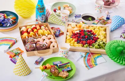 Ein bunter Partytisch mit Obstplatten, Muffins, Donuts, Süßigkeiten, Getränken und festlichen Accessoires wie Hüten und Servietten auf einer weißen Tischdecke.
