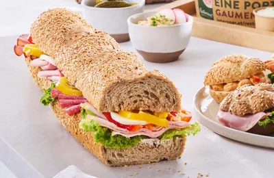 Ein Sesam-Baguette-Sandwich, gefüllt mit Salat, Schinken, Paprika und Radieschen. Im Hintergrund liegen kleine Brötchen auf einer weißen Unterlage.