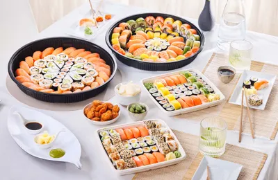Verschiedene Sushi-Platten mit Nigiri, Rollen, Sojasauce und Gewürzen auf einer weißen Tischdecke, dazu Getränke und Essstäbchen.