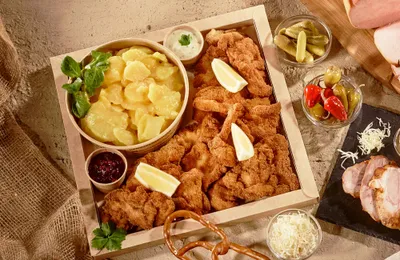 Eine Platte mit Backhendl, Kartoffelsalat, Zitronenschnitzen, Brezeln und verschiedenen eingelegten Gemüsesorten, dazu Dips und Beilagen.
