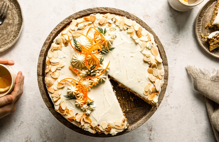 Karottenkuchen mit Frischkäseglasur, Mandelscheiben und Karottengarnitur auf einem Holzständer. Ein Stück fehlt, sodass man das Innere sehen kann.
