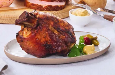 Gebratene Schweinshaxe auf einem Teller mit Senf, Gewürzgurken und Petersilie, serviert auf einer weißen Tischdecke, im Hintergrund Brot.