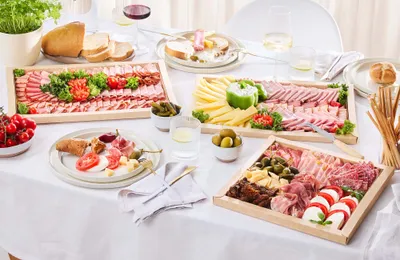 Ein gedeckter Tisch mit Wurst- und Käseplatten, auf denen Fleisch, Käse, Oliven, Tomaten, Brot und Getränke auf einer weißen Tischdecke angerichtet sind.