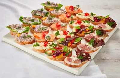 Eine Auswahl an offenen Sandwiches mit Räucherfisch, Fleisch, Gemüse und Kräutern auf einem weißen Tablett, serviert auf einer hellen Tischdecke.