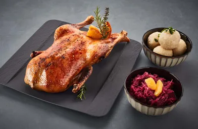 Gebratene Ente, garniert mit Kräutern und Orangenscheiben, auf einer schwarzen Platte, dazu Knödel und Rotkohl.