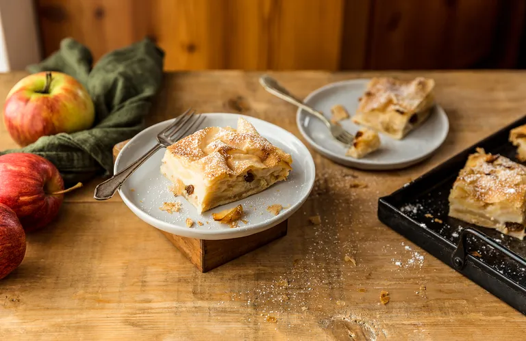 Stücke Apfelstrudel mit Rosinen auf Tellern, umgeben von frischen Äpfeln und einer Pfanne, auf einem rustikalen Holztisch.