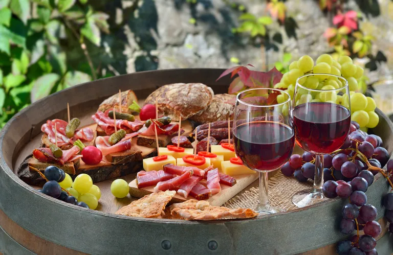 Ein rustikales Wein- und Wurstplatten-Buffet auf einem Holzfass mit Käse, Wurstwaren, Brot, Weintrauben und zwei Gläsern Rotwein im Freien.