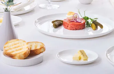 Elegante Tischdekoration mit Rindertartar, garniert mit Gewürzgurken, Zwiebeln, Microgreens und einem halbierten gekochten Ei; dazu geröstetes Brot und Butter.