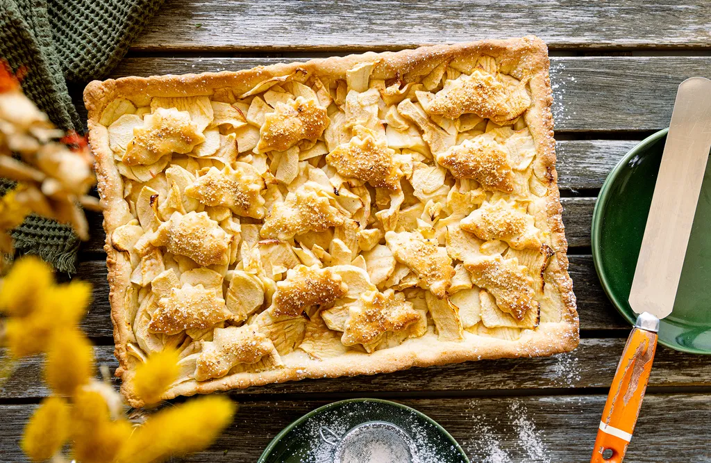 Ein rechteckiger Apfelkuchen mit blattförmigem Teig auf einem rustikalen Holztisch, daneben ein grüner Teller und ein Wellenschliffmesser.
