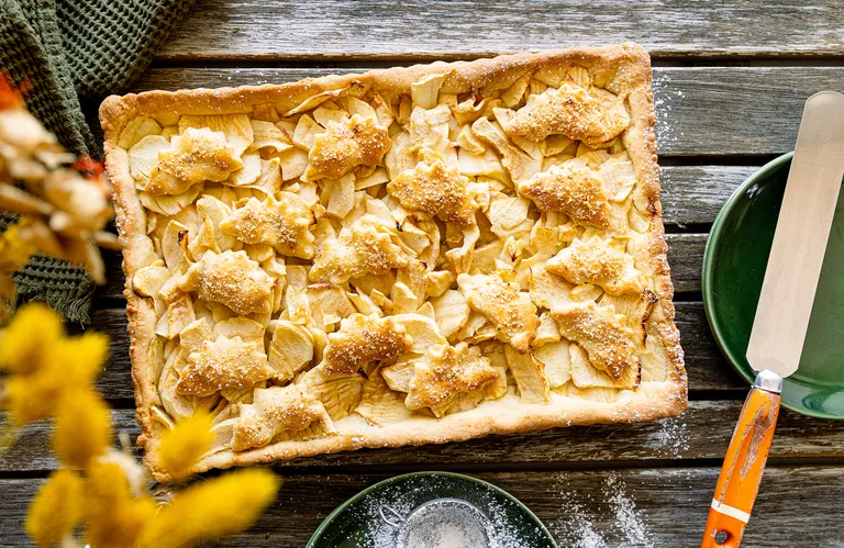 Ein rechteckiger Apfelkuchen mit blattförmigem Teig auf einem rustikalen Holztisch, daneben ein grüner Teller und ein Wellenschliffmesser.