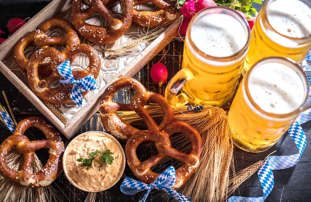Brezeln mit einer blau-weißen Schleife, zwei Krüge Bier, Weizenhalme, Radieschen und eine Schüssel mit Dip auf einem rustikalen Holztisch.