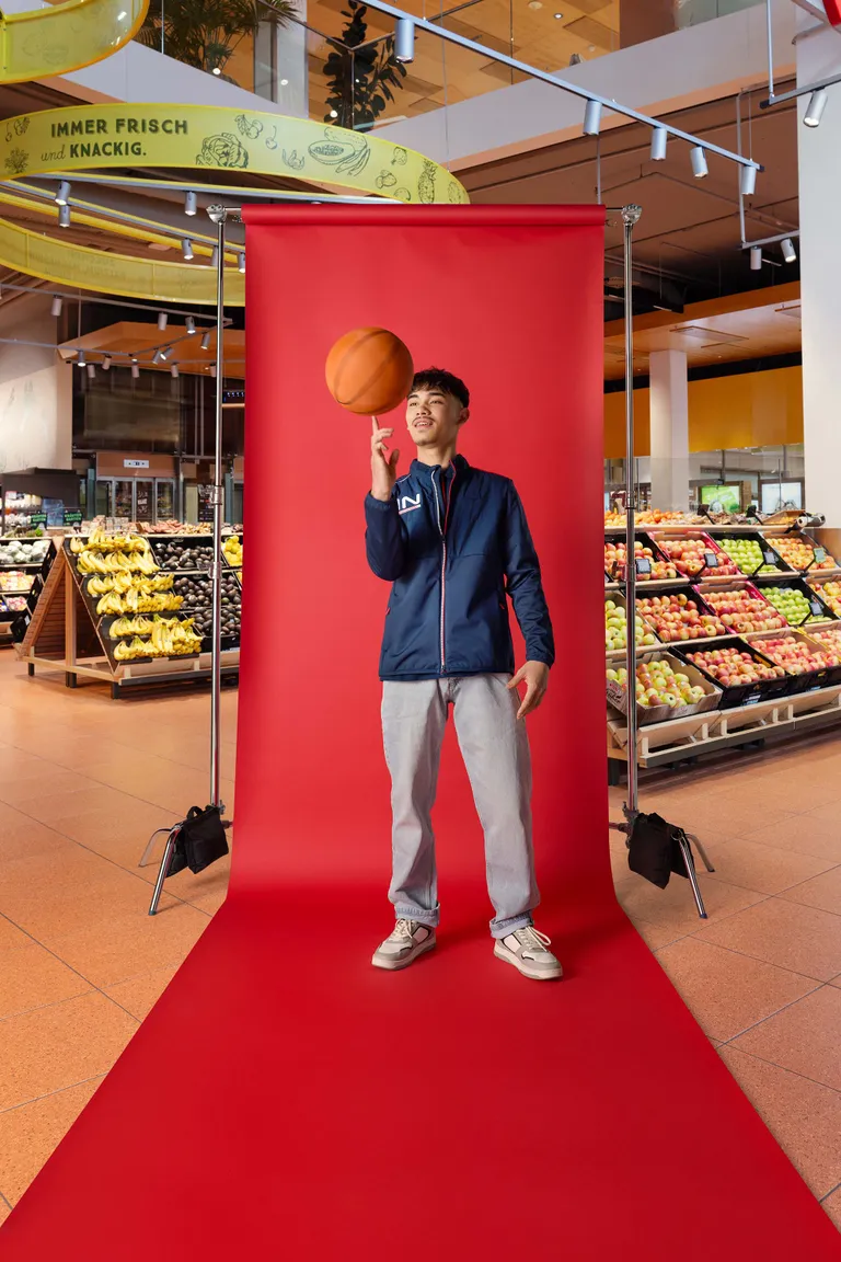 Lehrling lässt einen Basketball auf dem Finger kreisen und steht vor rotem Fotohintergrund im Markt