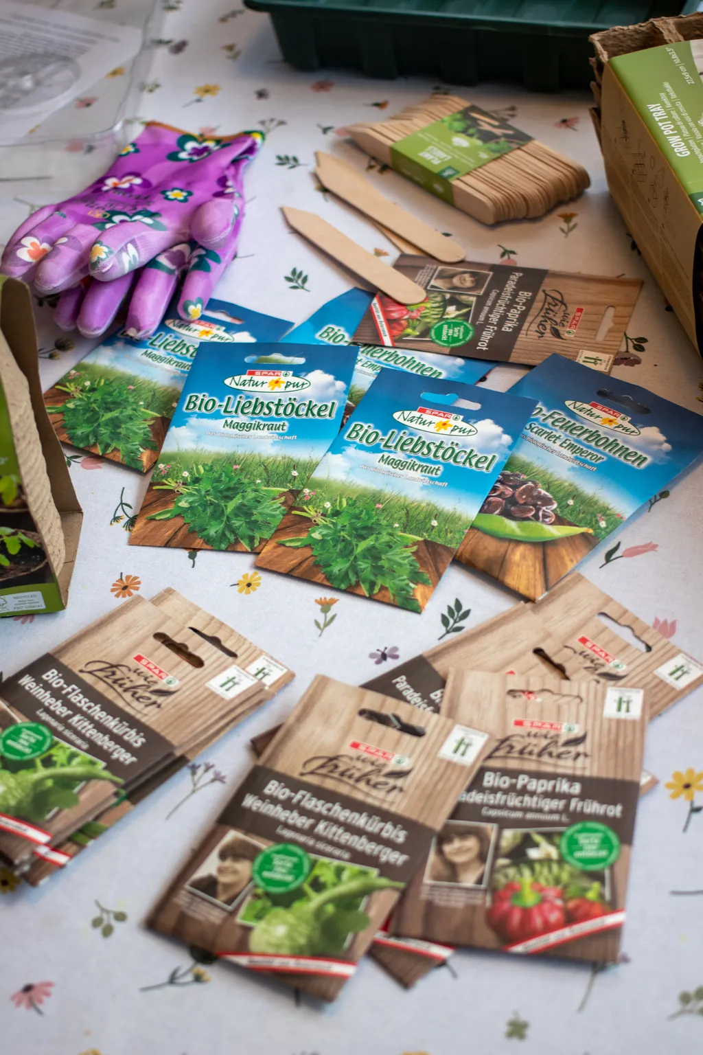 Samen, Saatschalen und Gartenhandschuhe auf einer geblümten Tischdecke
