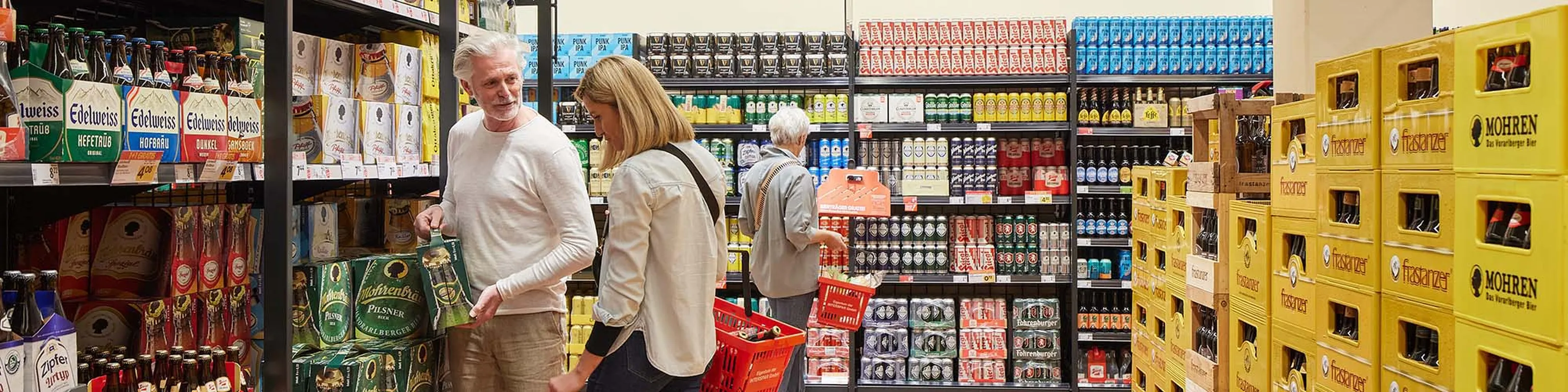 Menschen beim Einkaufen in einem hell erleuchteten Laden, umgeben von Regalen voller verschiedener Biermarken und gelben Kisten auf der rechten Seite.
