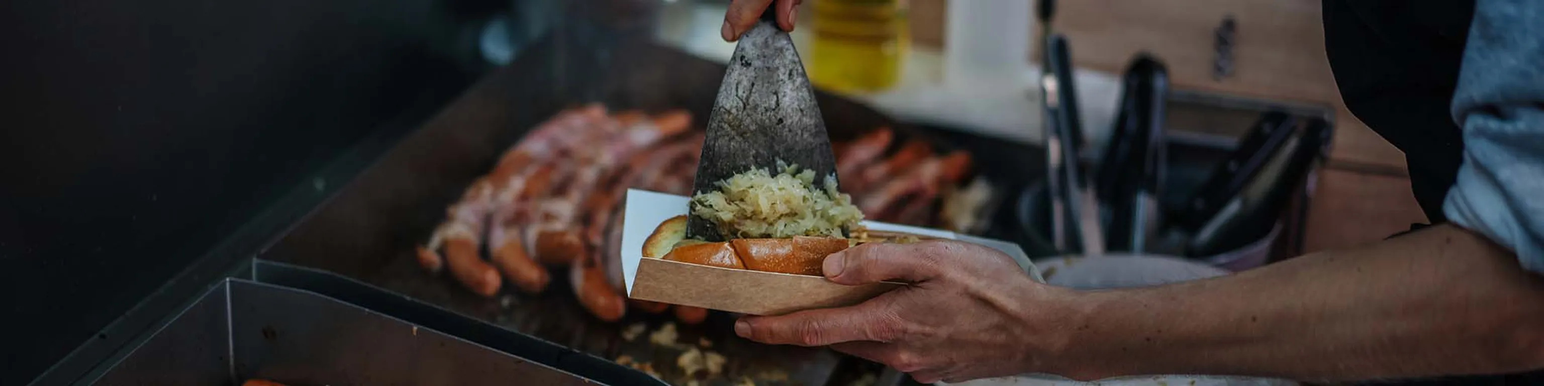 Eine Person bereitet eine Wurst im Brötchen zu und gibt Sauerkraut mit einem Pfannenwender darauf, während im Hintergrund weitere Würste auf dem Grill braten.