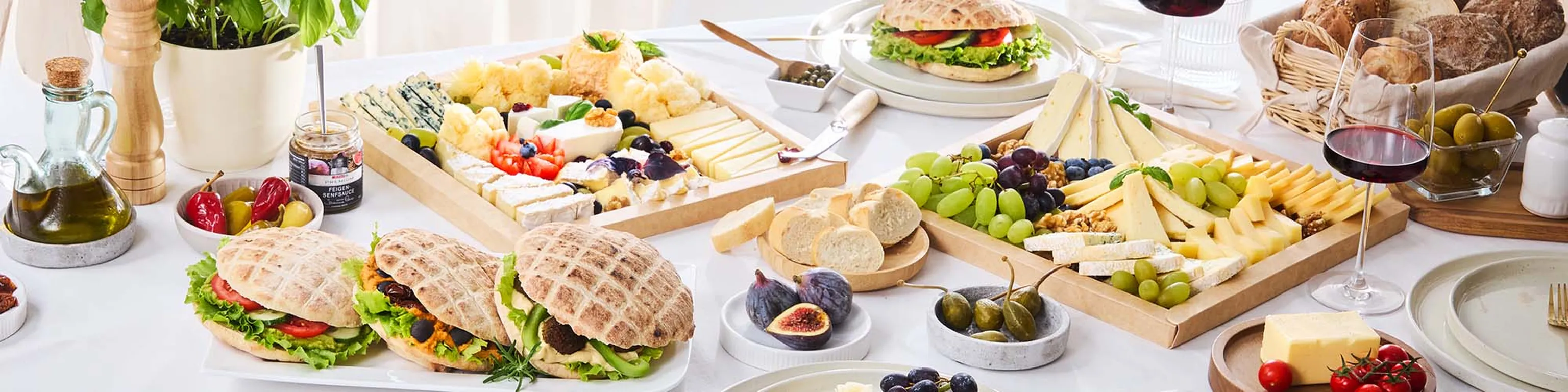Eine elegante Tischdekoration mit Sandwiches, verschiedenen Käsesorten, Weintrauben, Feigen, Brot und Rotwein auf einem Tisch mit dekorativen Elementen.