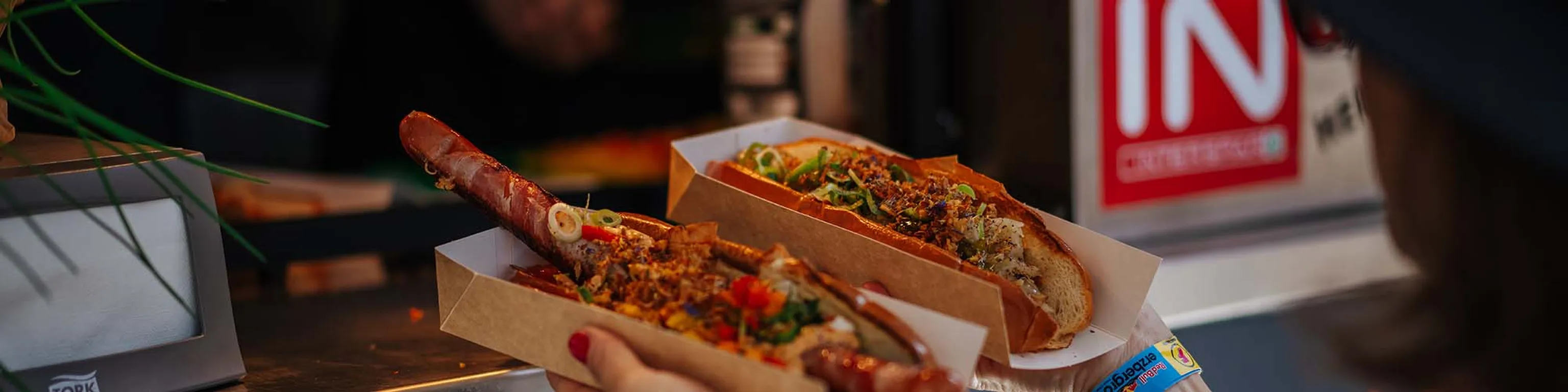 Zwei große Hotdogs in Pappschalen mit verschiedenen bunten Belägen, die eine Person vor einem Imbissstand in der Hand hält.