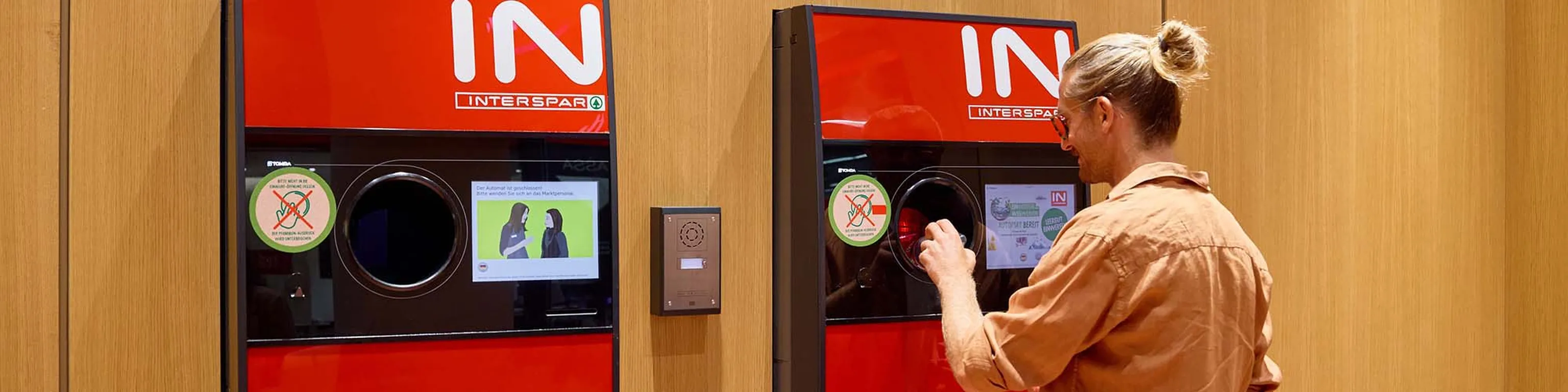 Eine Person, die einen Flaschenrücknahmeautomaten INTERSPAR benutzt, vor einer Holzwand und mit Anleitungsaufklebern am Automaten.