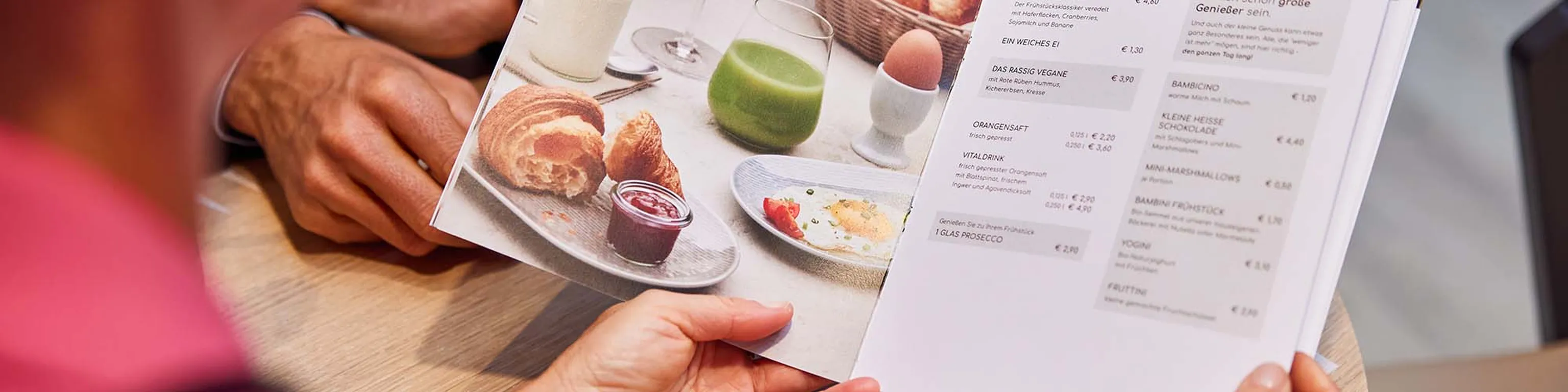 Hände, die eine Speisekarte mit Frühstücksangeboten und Preisen halten, darauf ein Foto von einem Croissant, Marmelade, einem grünen Saft und einem weichgekochten Ei.